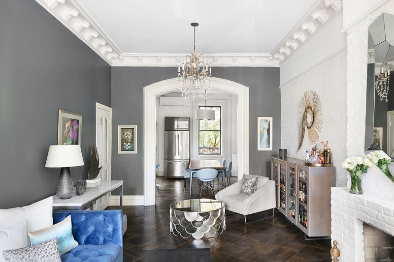 A living room area with hardwood floors, several couches, a chandelier, dark grey walls, and exposed white brick.