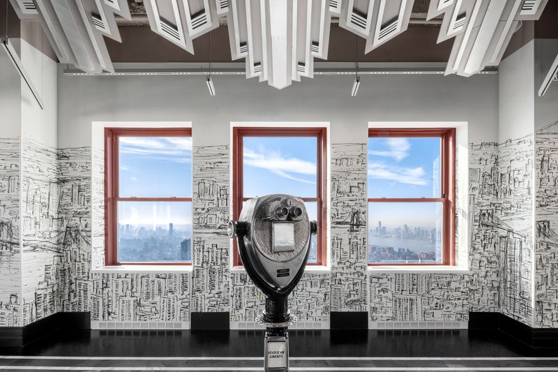 Three windows with panoramic views of the Manhattan skyline, a binocular, and a wall covered in a black and white drawing.