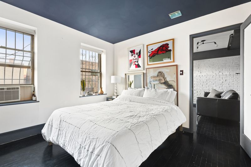 A bedroom with a medium-sized bed, two windows, hardwood floors, and several paintings hanging on the wall. 
