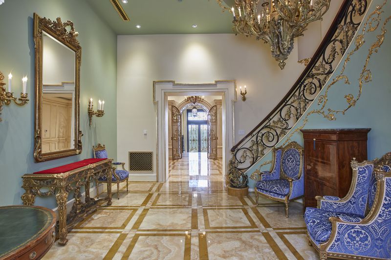 A foyer with marble floors, a staircase with elegant railings, and two blue chairs.