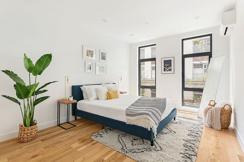 A bedroom with a small bed, a planter, hardwood floors, and two floor-to-ceiling windows.