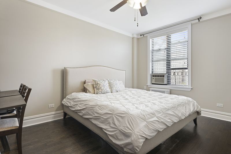A bedroom with hardwood floors a large bed, a window, beige walls, and crown moldings. 