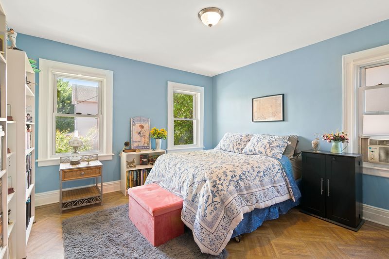 A bedroom with a medium-sized bed, light blue walls, three windows, base moldings, and hardwood floors. 