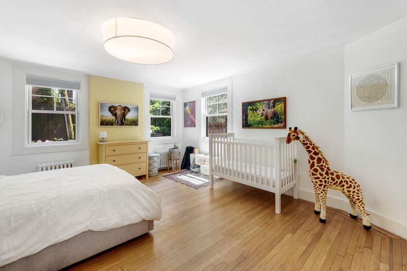 A bedroom with a crib, a toy giraffe, hardwood floors, a bed, and three windows.