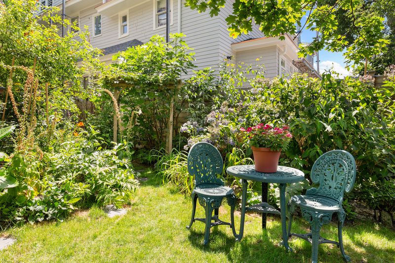 A landscaped backyard filled with plants and a round green table with two chairs.