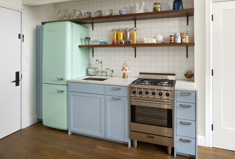 A small kitchen with a light green fridge, light blue cabinetry, and white tiles on its walls.