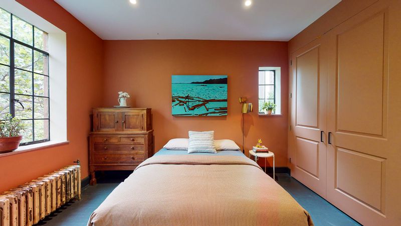 A bedroom with dark orange walls, a bed, and a large casement window.