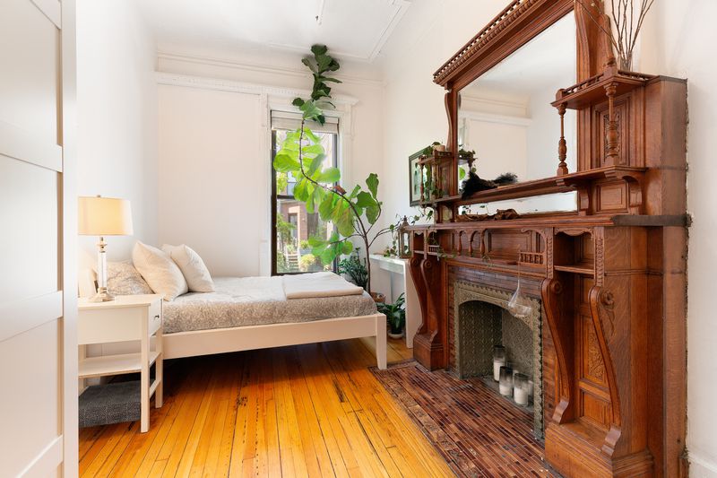 A room with a white bed, large houseplant, and huge wooden fireplace.